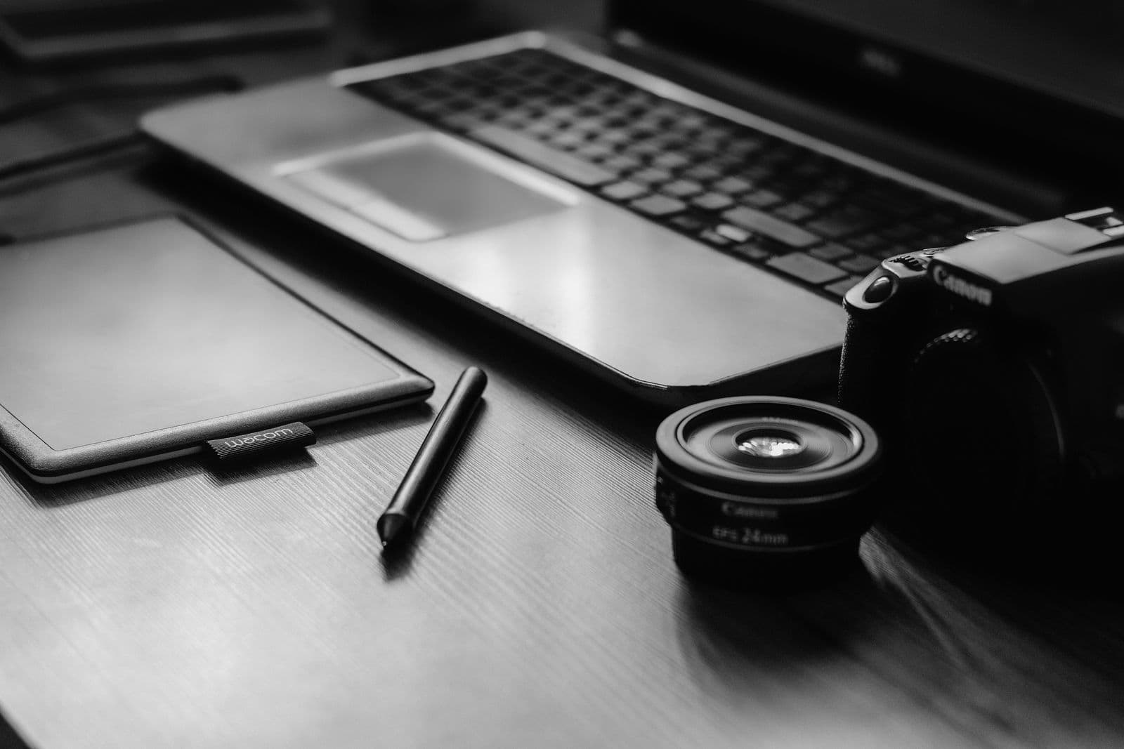 Monochrome desk with a camera, lens, laptop, and tablet